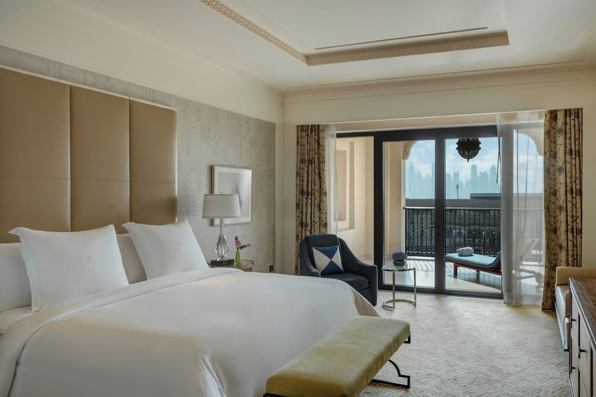 A hotel room with a large bed, a chair, a small table, and a balcony with a city view.