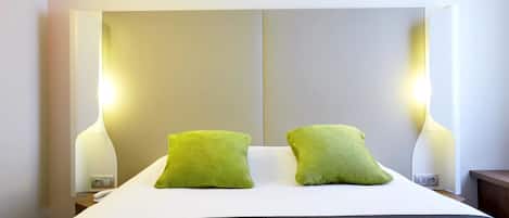 Una cama bien hecha con dos almohadas verdes, un edredón blanco y una manta oscura. Hay dos lámparas de noche de diseño moderno, y una mesita de noche con un teléfono y un enchufe.