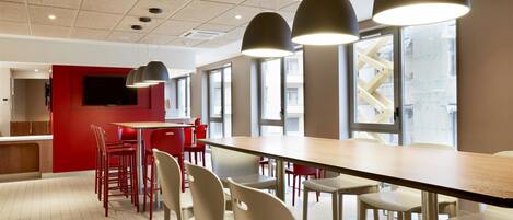 A modern restaurant with a long wooden table, red chairs, and pendant lights.