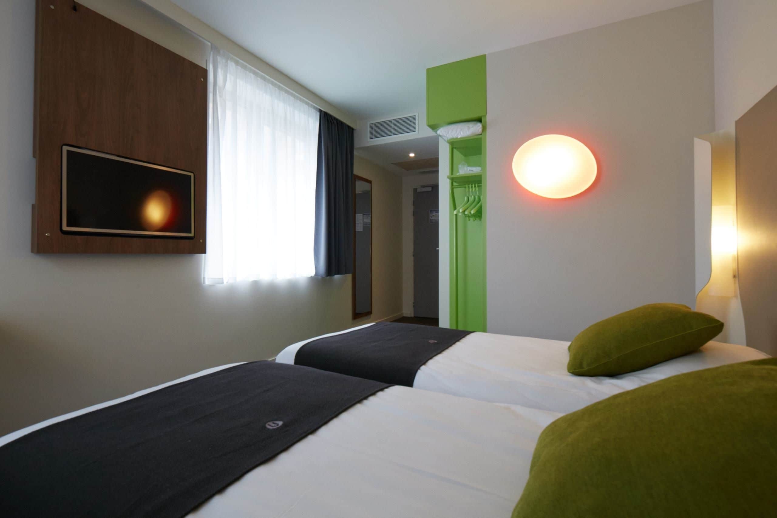 A hotel room with two beds, a television, a green wall, and a round wall light.