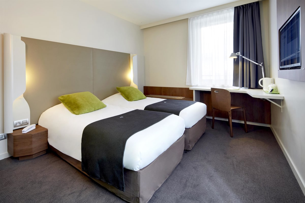 A hotel room with two beds, a desk, a chair, a nightstand, and a window with curtains.