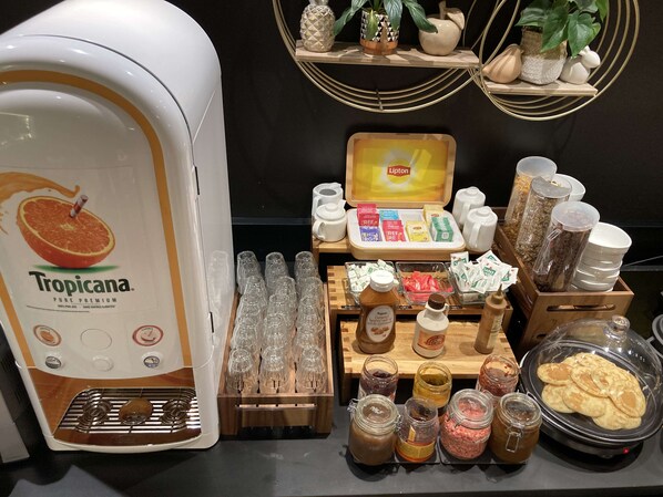 Estación de desayuno con dispensador de jugo Tropicana, cristalería, condimentos y una olla de cocción lenta con panqueques.