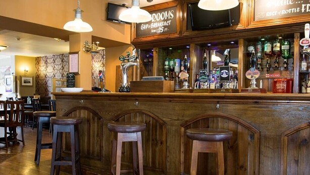 A bar area with wooden bar stools, a counter with a variety of bottles, and a sign indicating a breakfast buffet. - The Albert (Scarborough)