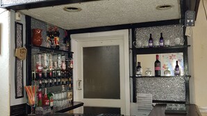 Una zona de bar con estanterías llenas de botellas, cuadros en la pared y una botella de Coca-Cola en la barra.