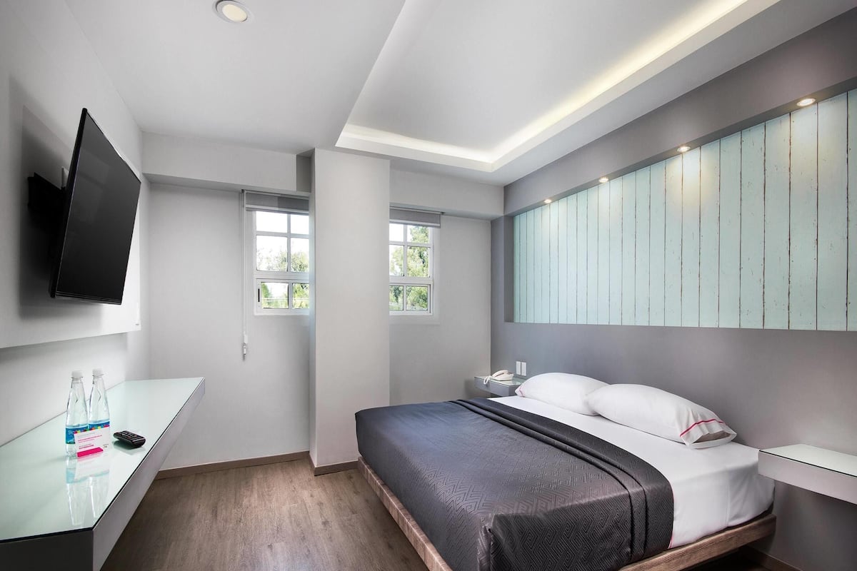 Una habitación de hotel moderna con una cama, mesita de noche, televisor y un ventanal grande.