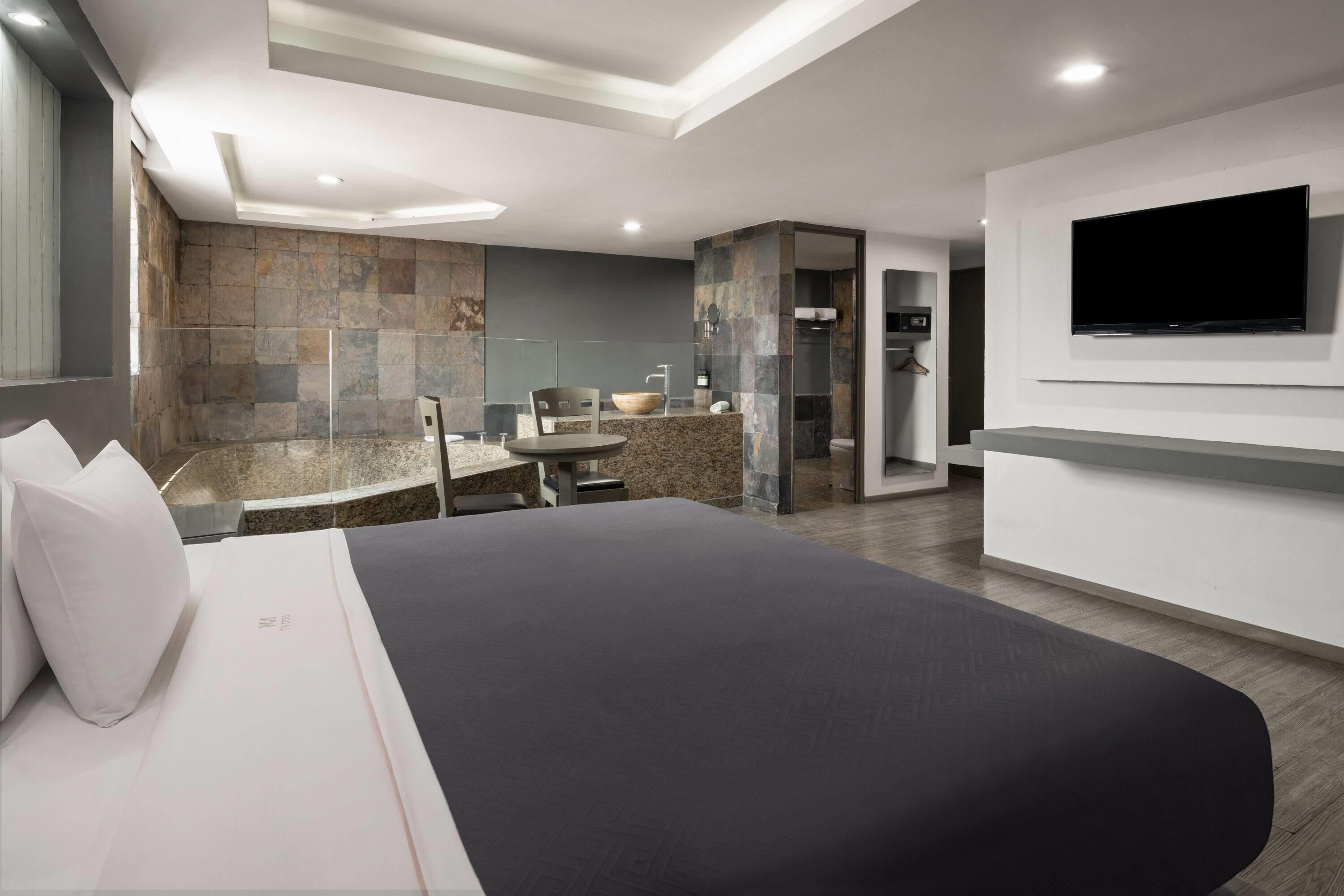 A modern hotel room with a large bed, a seating area with a table, a TV mounted on the wall, and a stone wall.