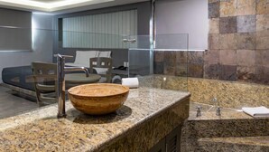 Un baño moderno con una amplia encimera de granito, un lavabo de madera, cabina de ducha de vidrio y una zona de estar con una silla y una mesa.