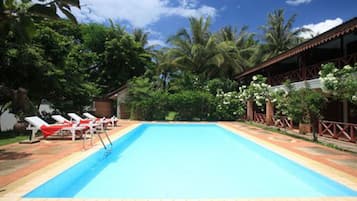 A rectangular swimming pool with a tiled deck, surrounded by lush greenery and palm trees.