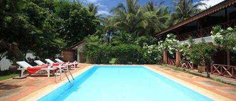Una piscina rectangular con solárium empedrado, rodeada de abundante vegetación y palmeras.