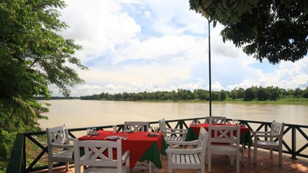 Un área de comedor al aire libre con sillas blancas y manteles rojos, con vista a un río.