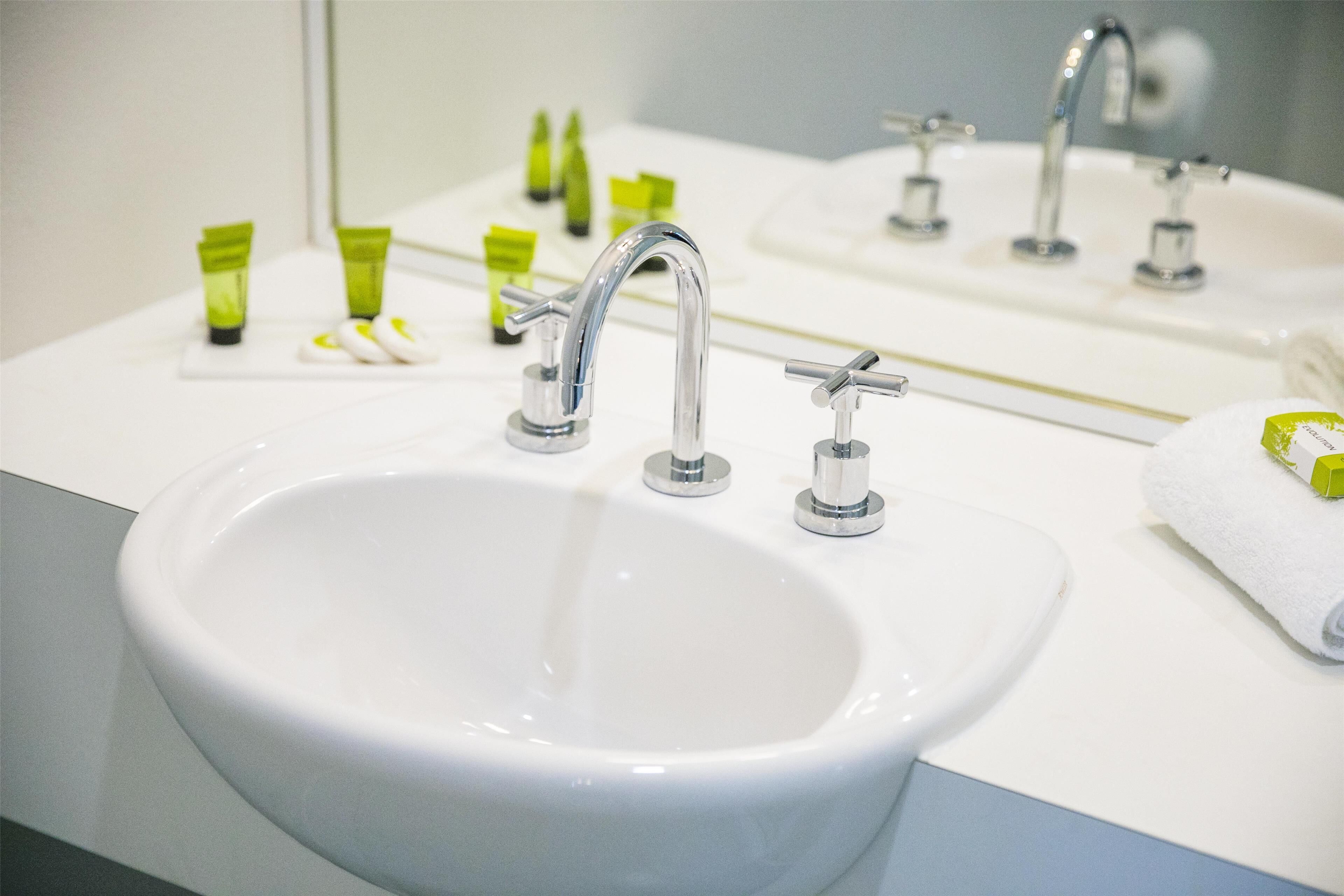 Un baño con un lavabo blanco, dos grifos y un espejo que refleja el lavabo y los grifos. Hay artículos de aseo verde y blanco sobre el mostrador del lavabo.