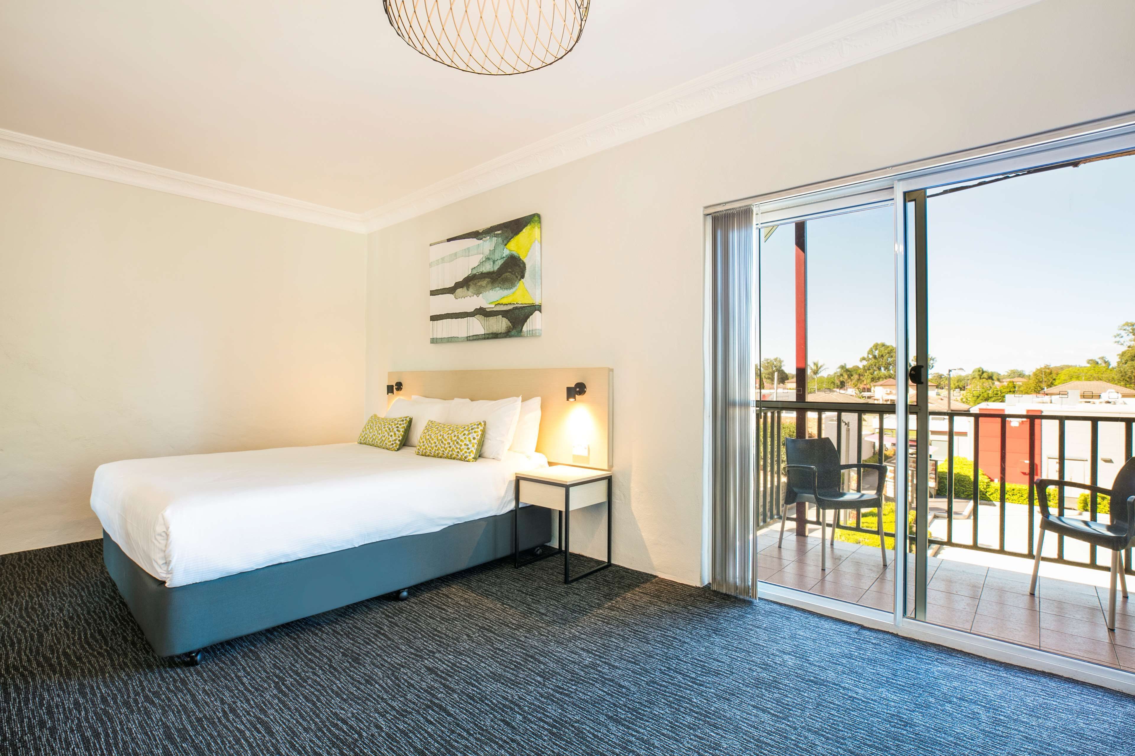 A modern hotel room with a large bed, a small bedside table, a wall-mounted artwork, and a balcony with chairs.