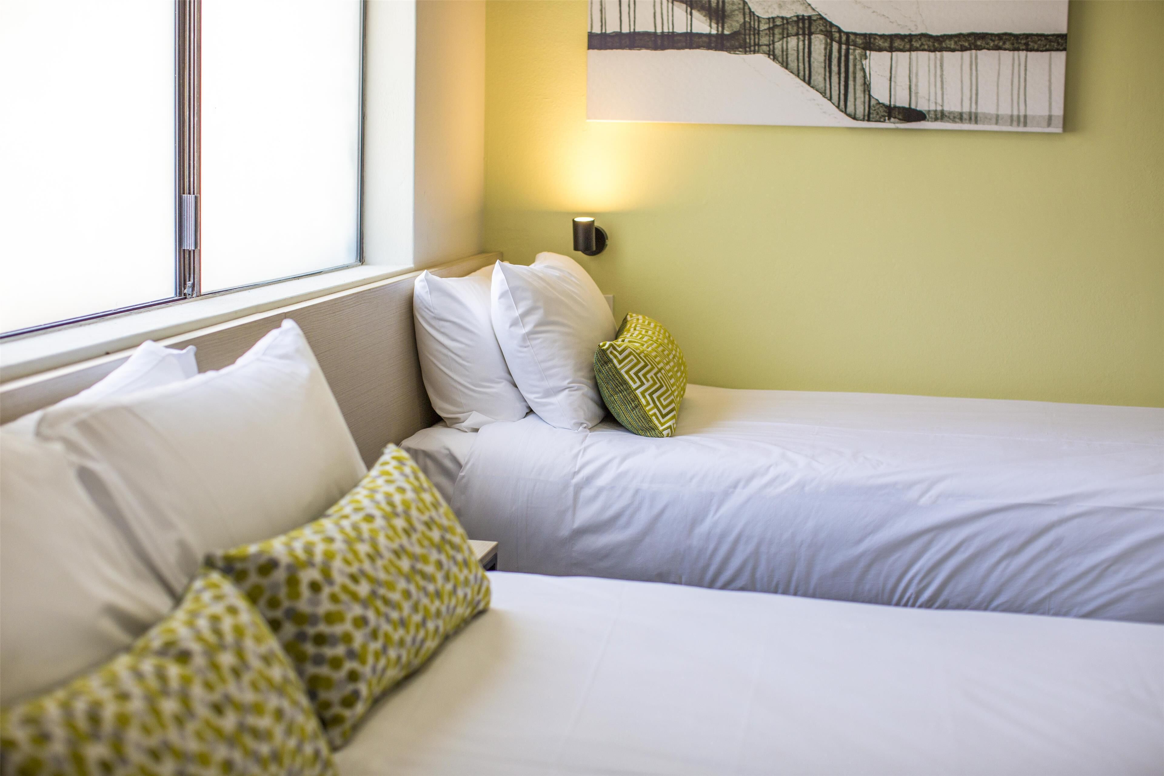 A hotel room with two beds, a window, and a wall-mounted light.
