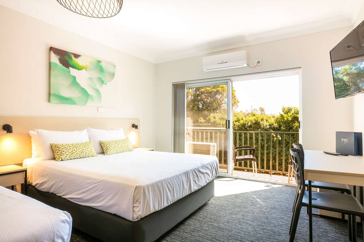 A hotel room with a large bed, a desk, and a balcony with a view of greenery.