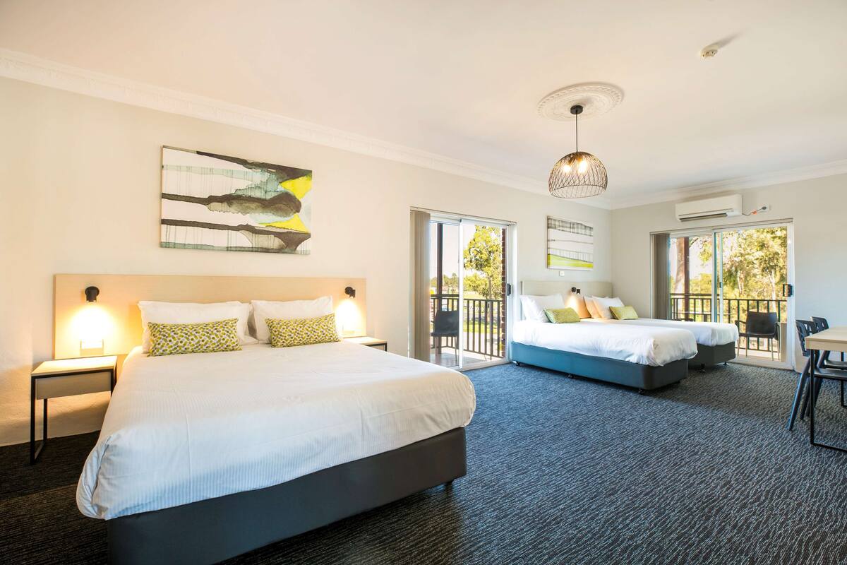A hotel room with a large bed, a smaller bed, a balcony with a table and chairs, and a ceiling light fixture.