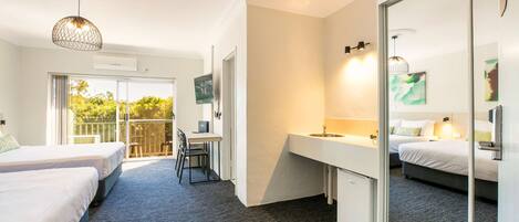 A hotel room with two beds, a desk, a large mirror, and a balcony with a view of greenery.