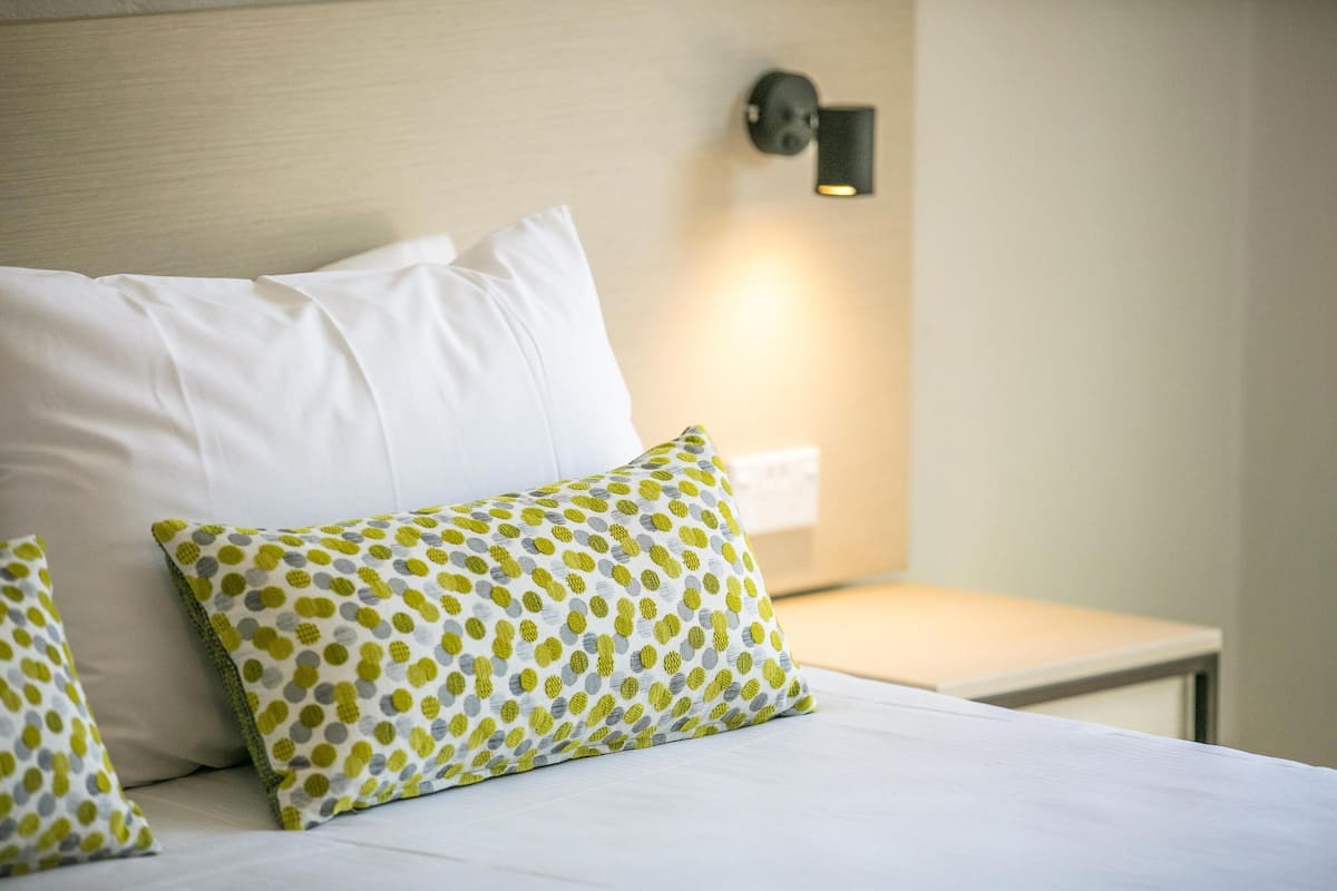 A neatly made bed with white and green patterned pillows, a bedside table, and a wall-mounted light fixture.