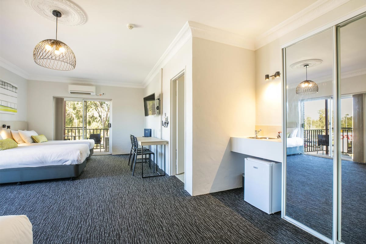 A hotel room with a bed, desk, and kitchenette. There is a large window and a balcony.