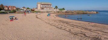 Uma praia com um edifício grande ao fundo, areal arenoso e pessoas praticando diversas atividades.
