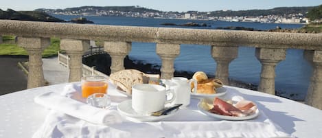 Mesa de desayuno con croissants, jamón y jugo de naranja, con vista a un pueblo costero.