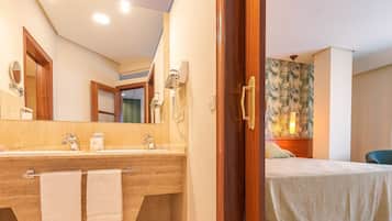 A hotel room with a wooden door, a large mirror, a double sink, and a bed with a light-colored bedspread.