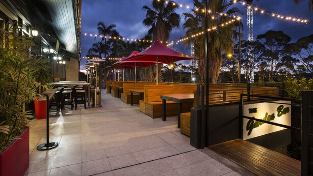 An outdoor dining area with wooden benches and red umbrellas, surrounded by palm trees and string lights. - Nightcap at the Ranch Hotel (Marsfield)