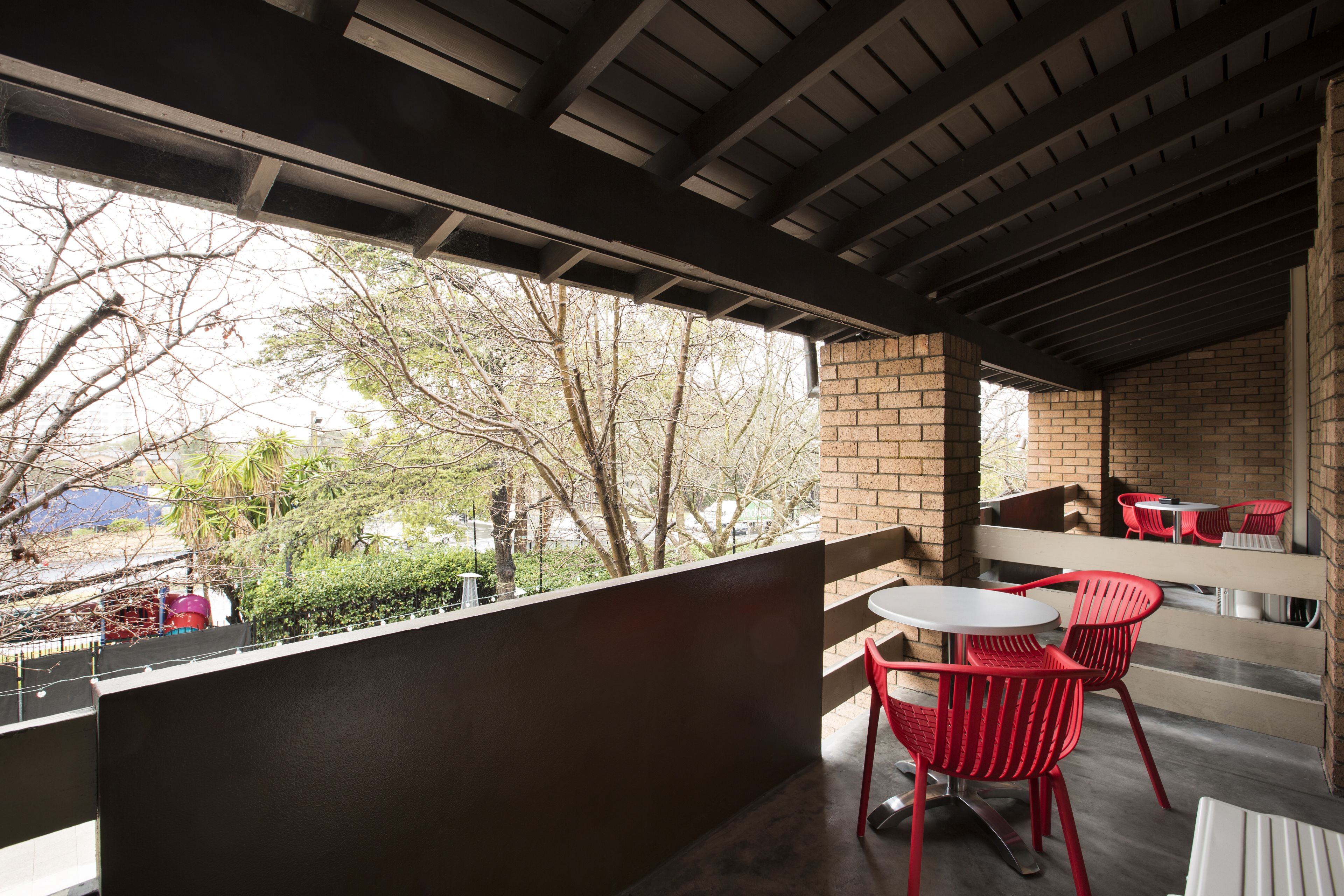 Un área de estar al aire libre cubierta con sillas rojas y una mesa blanca, con vistas a un jardín con árboles y un edificio al fondo.
