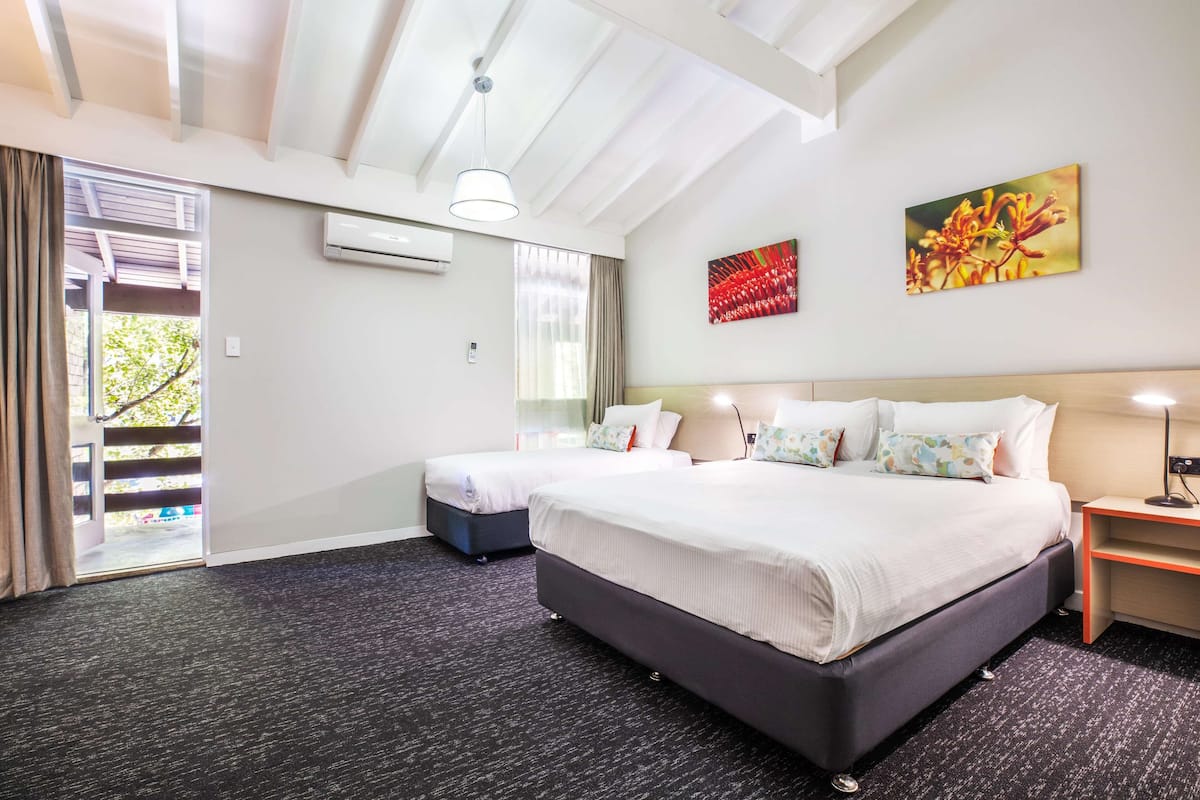 A hotel room with a large bed, a smaller bed, a desk, and a chair. There is a window with curtains, an air conditioning unit, and a painting on the wall.