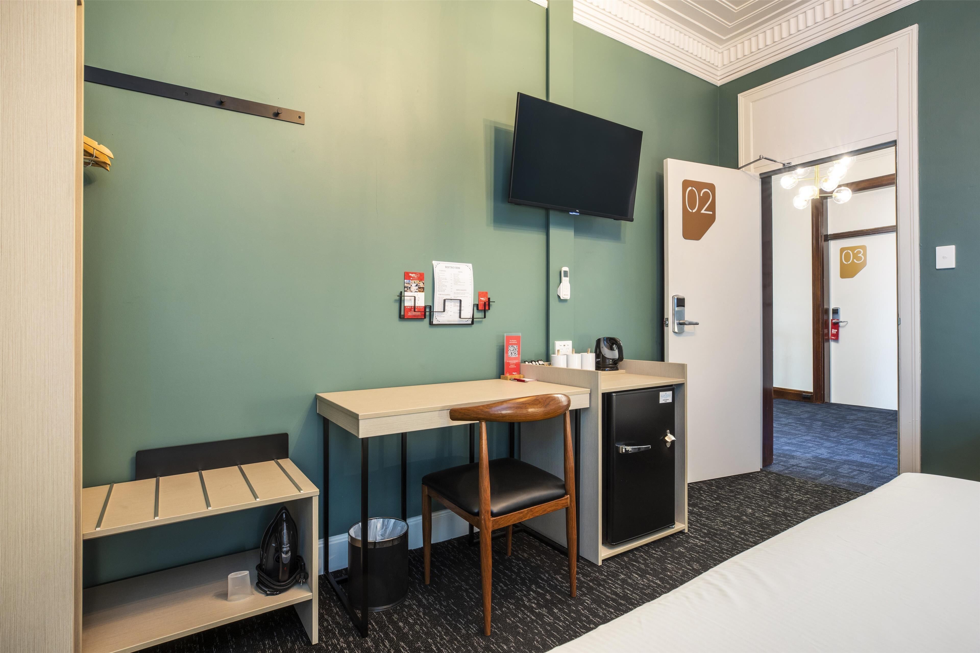 A hotel room with a bed, a desk, a chair, a TV, a minibar, and a door with room numbers.