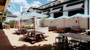 An outdoor patio area with picnic tables and striped umbrellas, surrounded by a building with a modern design. - Nightcap at Pritchards Hotel (Mt Pritchard)