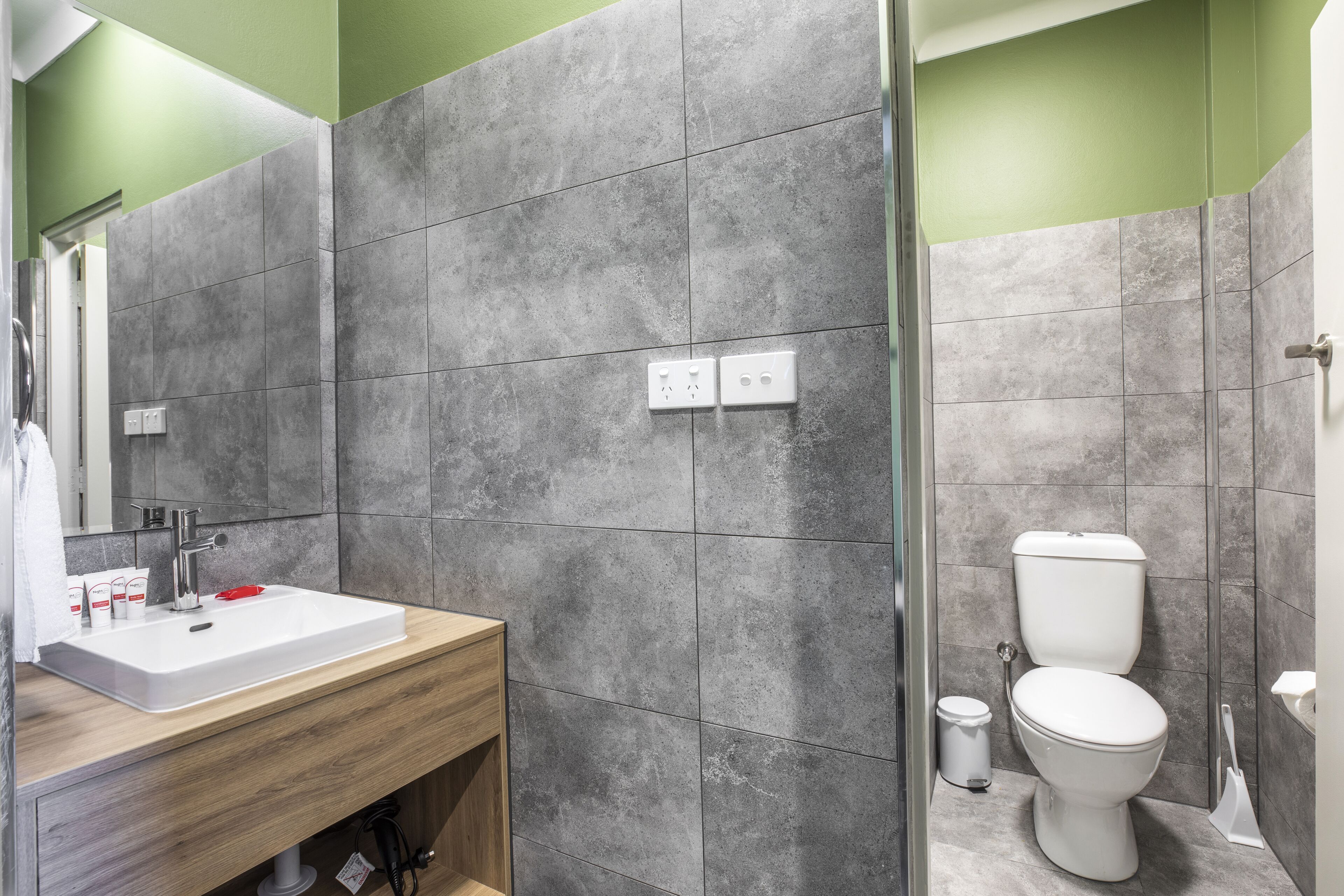 A bathroom with a wooden vanity, a white sink, a mirror, a toilet, and grey tiled walls.