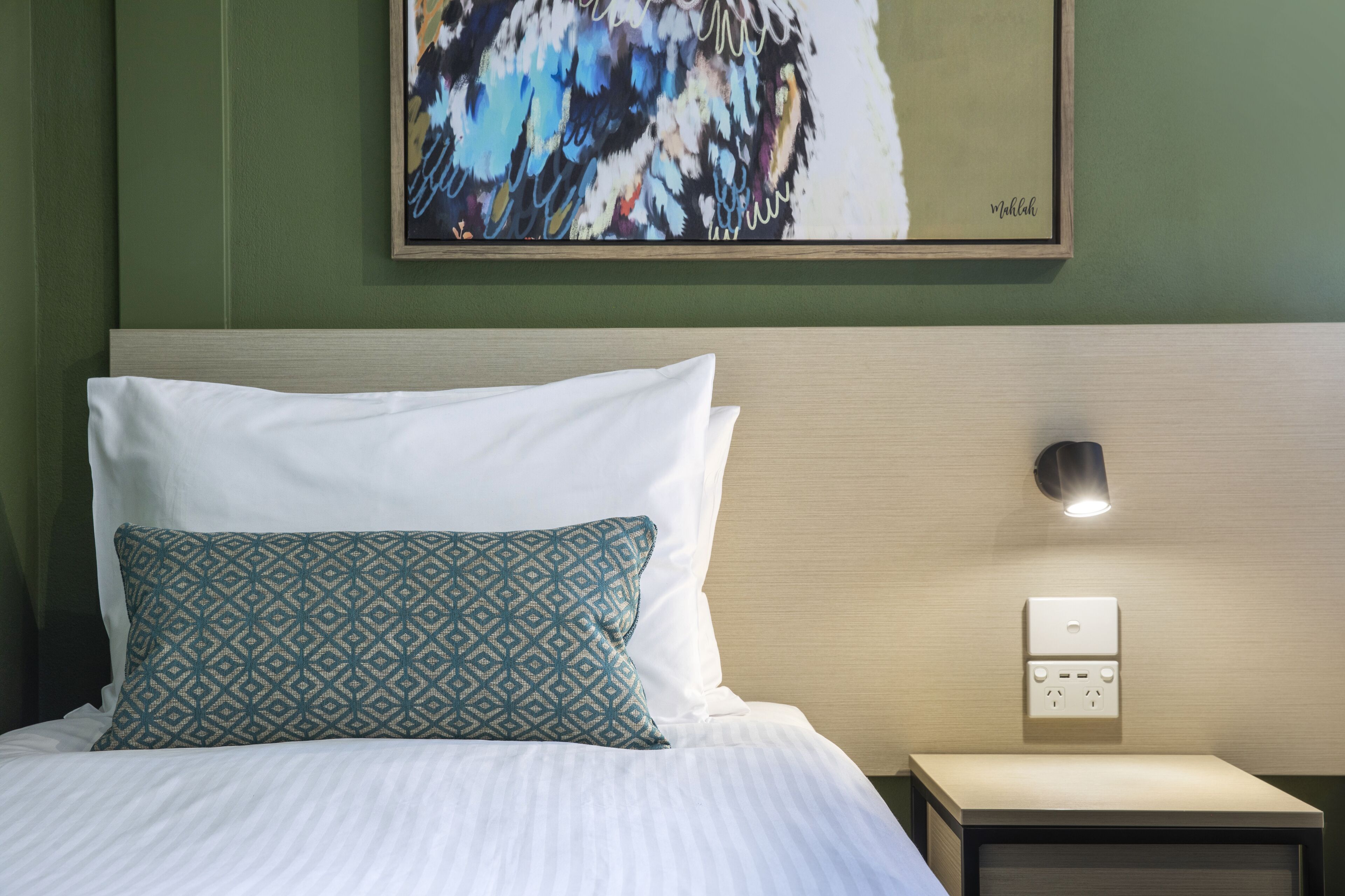 A neatly made bed with white and patterned pillows, a bedside table with a lamp, and a wall-mounted power outlet.