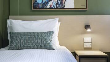 A neatly made bed with white and patterned pillows, a bedside table with a lamp, and a wall-mounted power outlet.