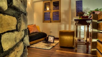 A cozy living room with a stone wall, a black sofa with cushions, a small ottoman, a wooden coffee table, and a window with wooden shutters.