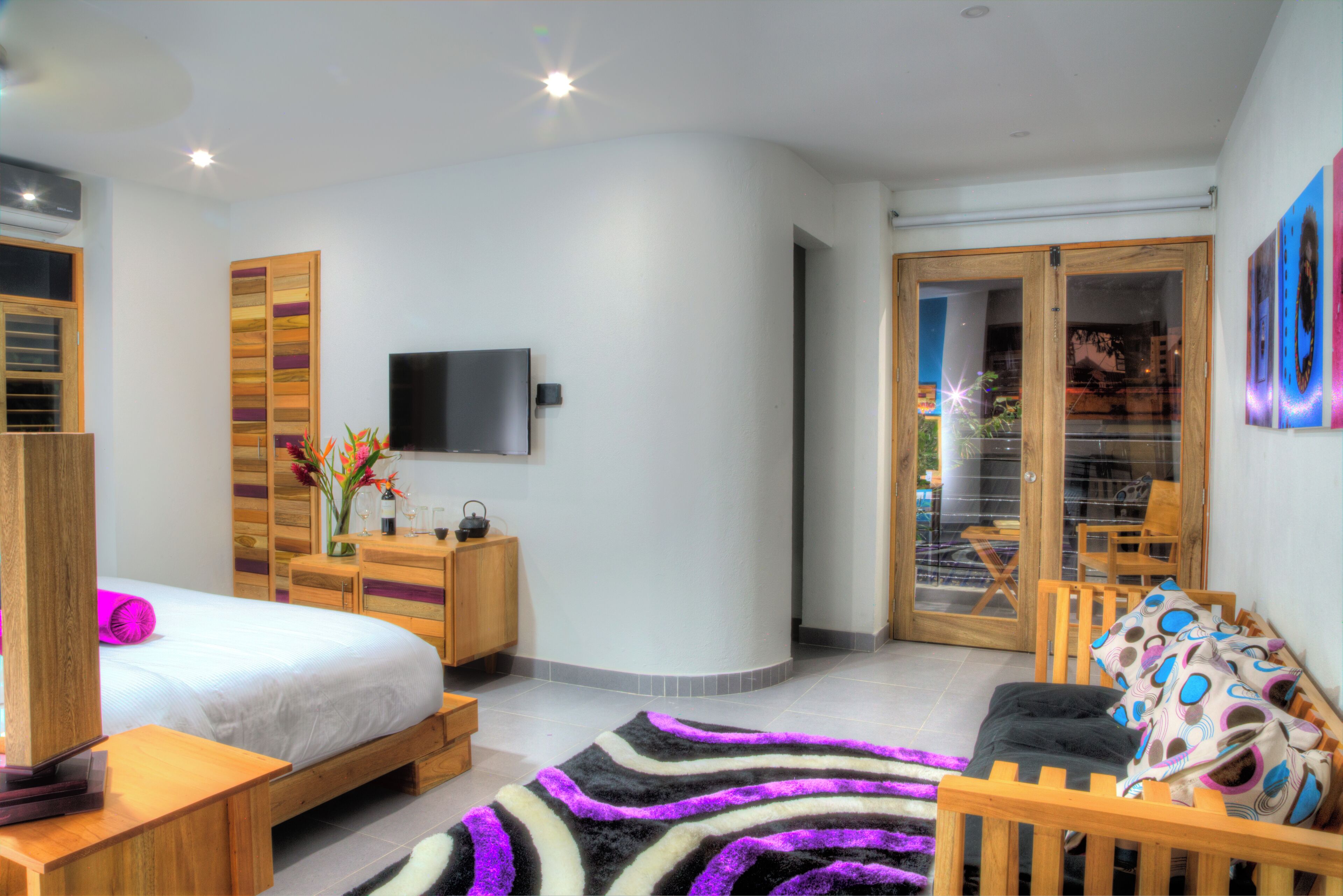 A modern bedroom with a bed, wooden furniture, a flat-screen TV, and a sliding glass door leading to a balcony.