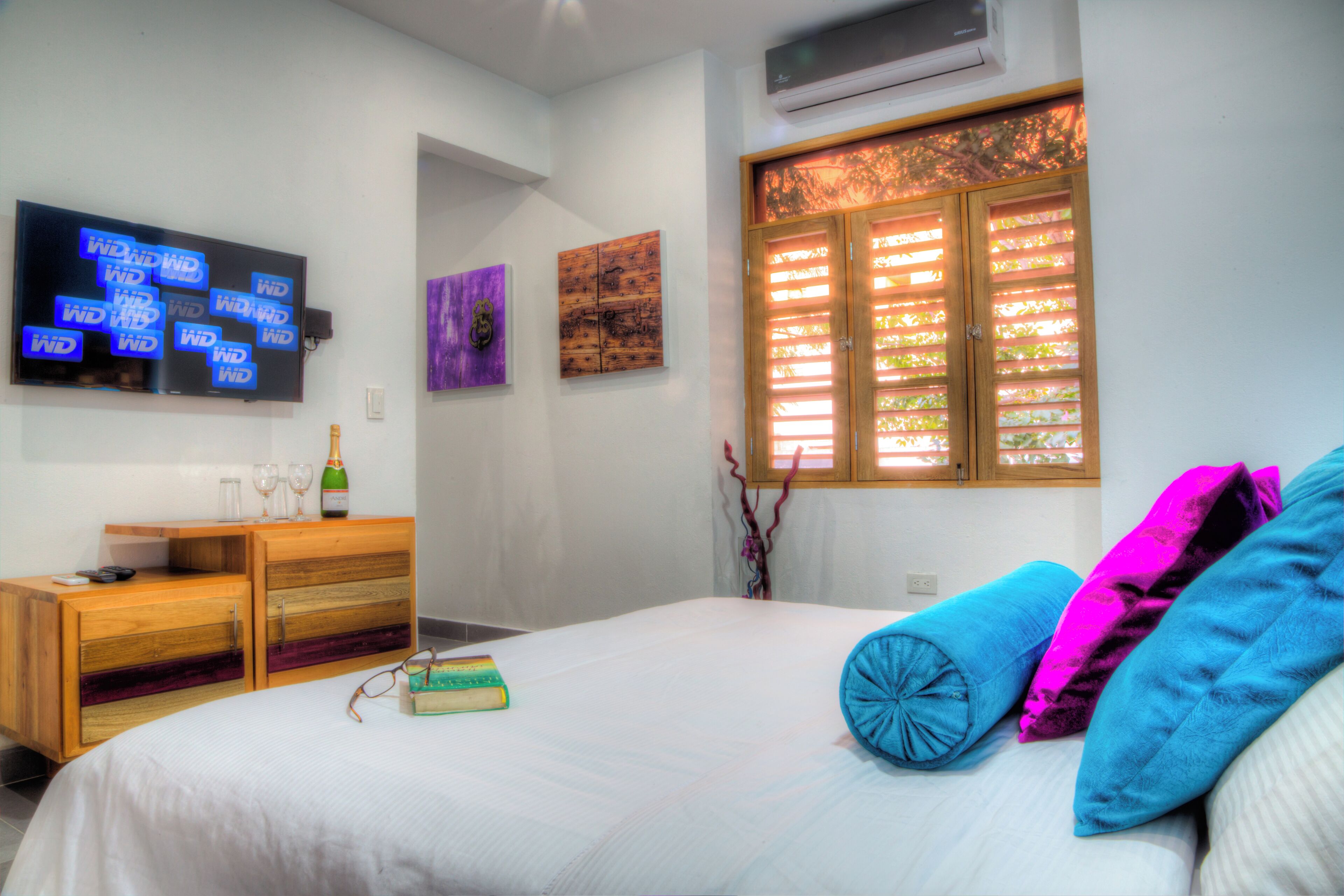 Une chambre à coucher avec un lit, un téléviseur fixé au mur, une commode en bois et une fenêtre avec des stores.