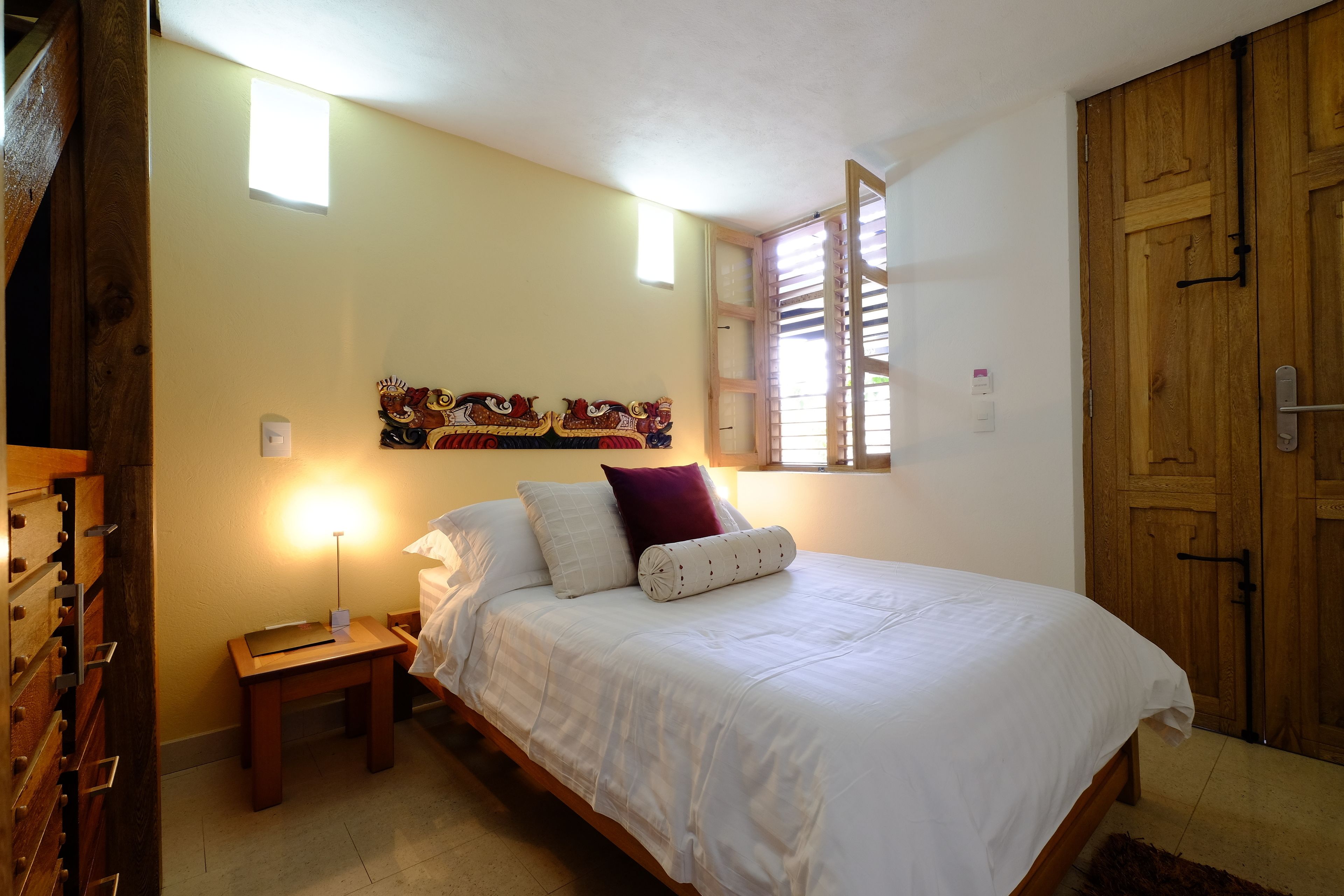 A bedroom with a bed, bedside table, lamp, artwork on the wall, and a wooden wardrobe.