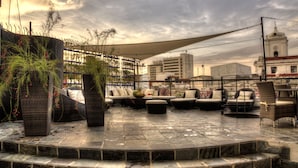 An outdoor seating area with wicker furniture and a stone floor, overlooking a cityscape with buildings and a church tower. - Casa de Leda, a Kali Hotel (Santa Marta)