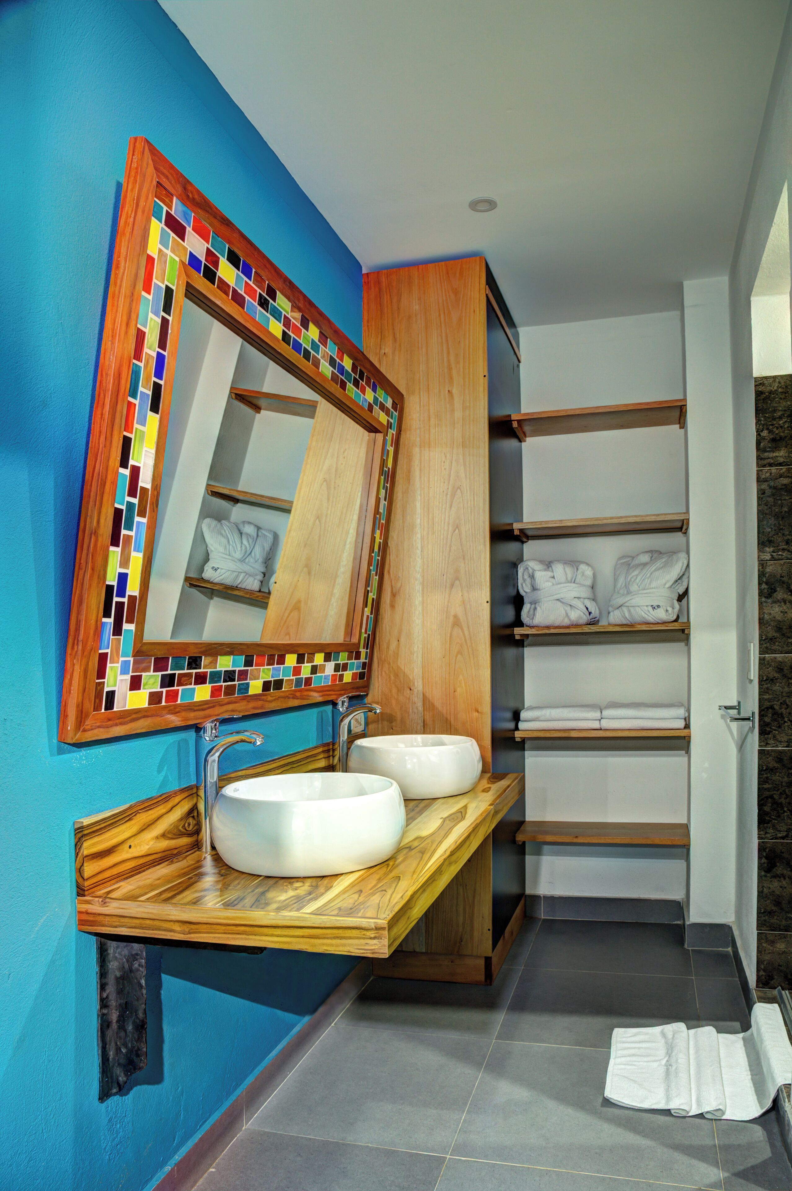 Un baño con tocador de madera, dos lavamanos, un espejo con marco de azulejos de colores y repisas con toallas.