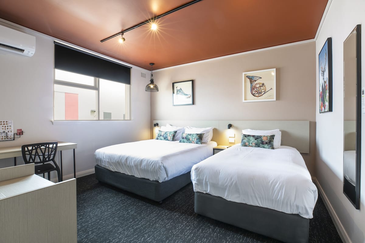 A hotel room with two beds, a desk, and a chair. There are wall-mounted lights and artwork.