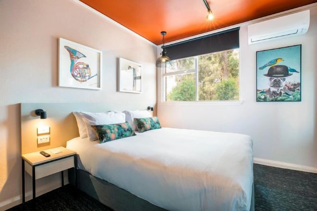 A bedroom with a bed, bedside table, wall art, and a window with a view of trees.