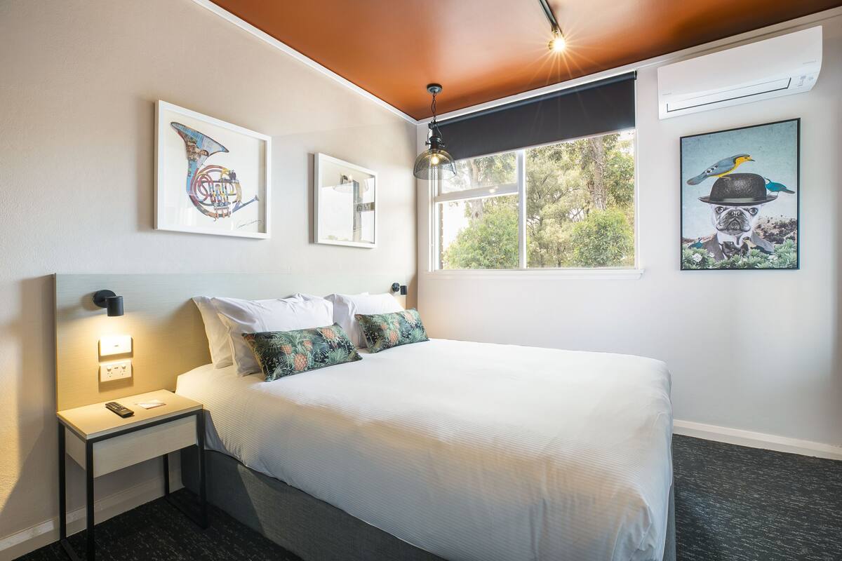 A neatly made bed with white linens and tropical patterned pillows, a bedside table with a remote control, and artwork on the walls.