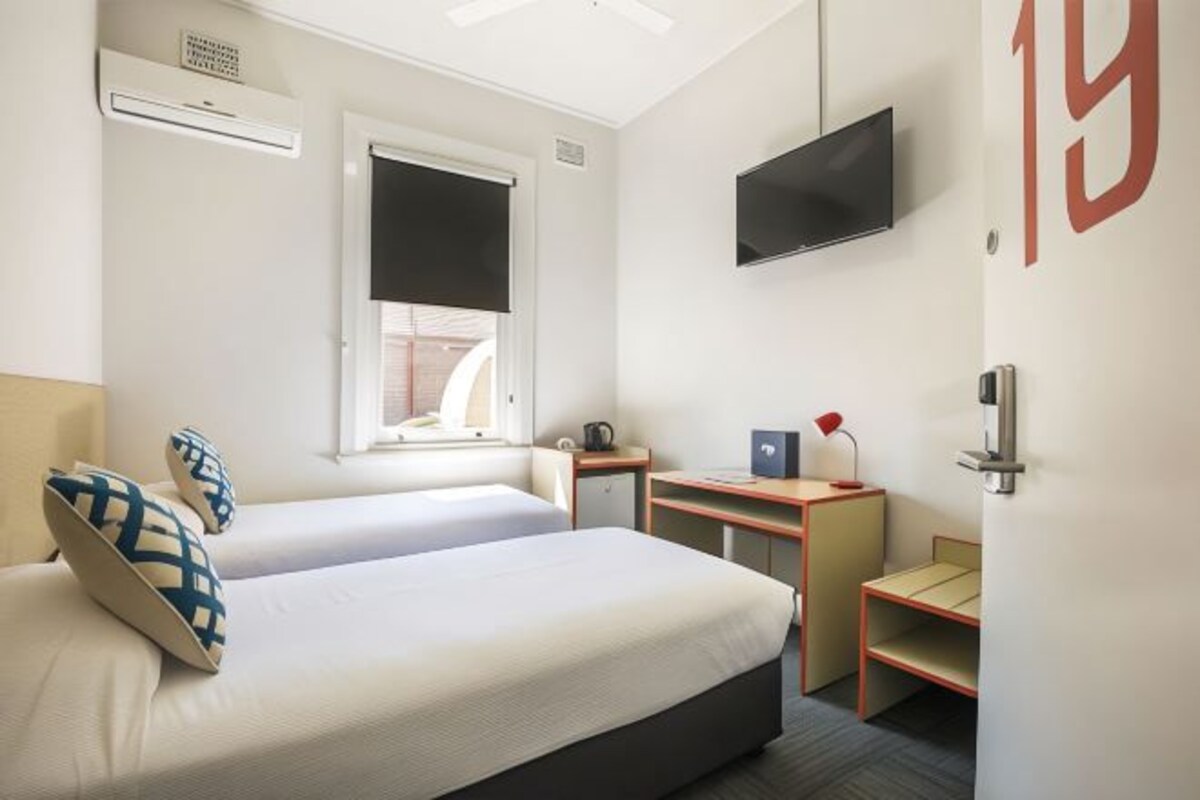 A hotel room with two beds, a desk, a TV, and a window with blinds.