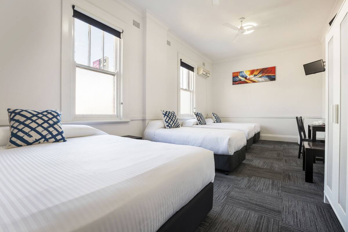 A hotel room with two beds, a desk, and a chair. There are windows with blinds, a ceiling fan, and a painting on the wall.