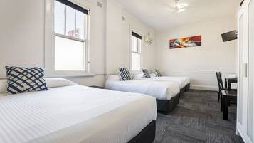 A hotel room with two beds, a desk, and a chair. There are windows with blinds, a ceiling fan, and a painting on the wall.