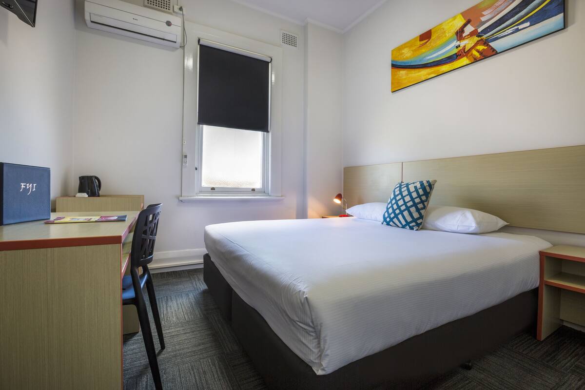 A hotel room with a bed, a desk, a chair, a window with a blind, a nightstand, a lamp, and a painting on the wall.