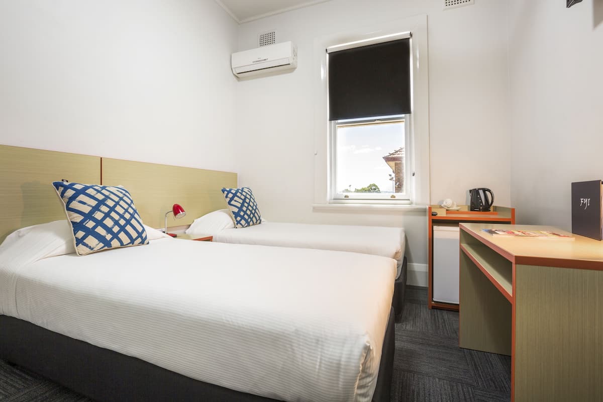 A hotel room with two beds, a desk, a small kitchenette, and a window with a view of a building outside.