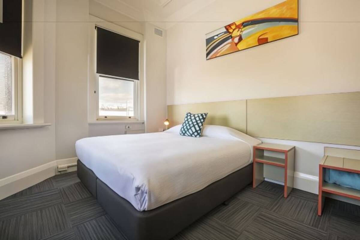 A hotel room with a large bed, a bedside table, a nightstand, a window with a view, and a painting on the wall.