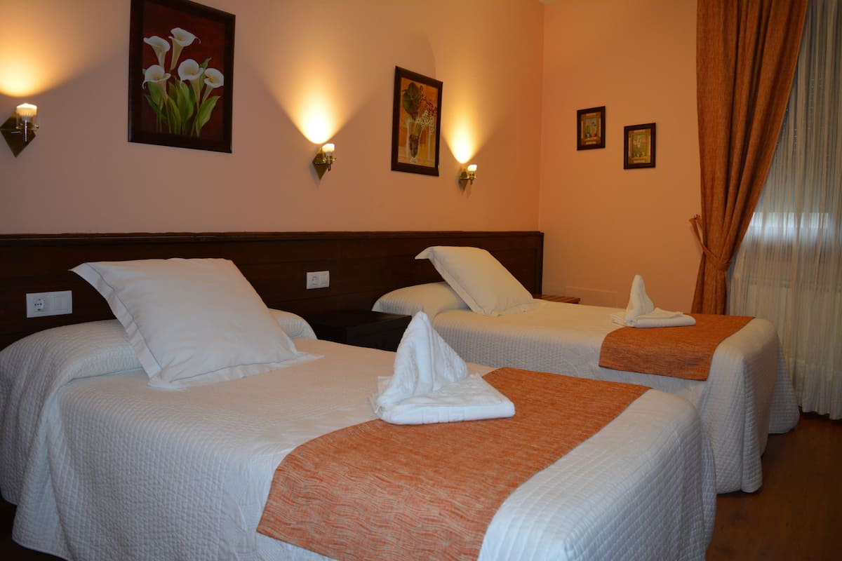 Quarto de hotel com duas camas, cada uma com roupa de cama branca e um cobertor marrom. Há luminárias fixadas na parede e quadros emoldurados.