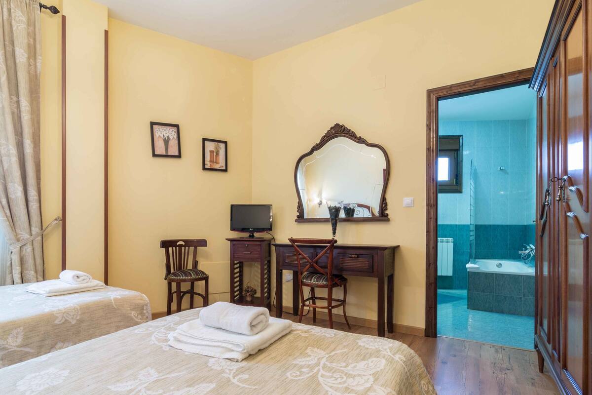Quarto de hotel com duas camas, uma secretária com televisão, um espelho e um banheiro visível por uma porta aberta.